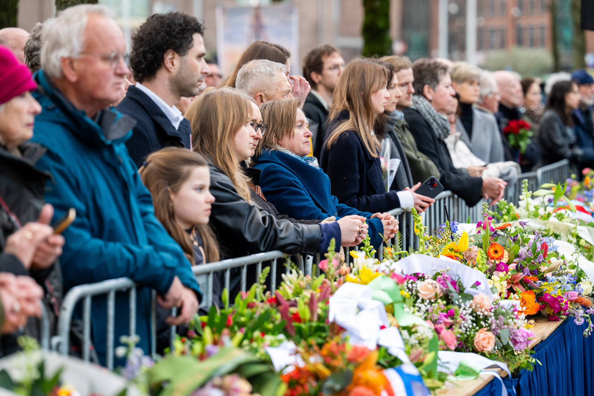 Tientallen mensen staan achter een hek, op de voorgrond liggen kransen