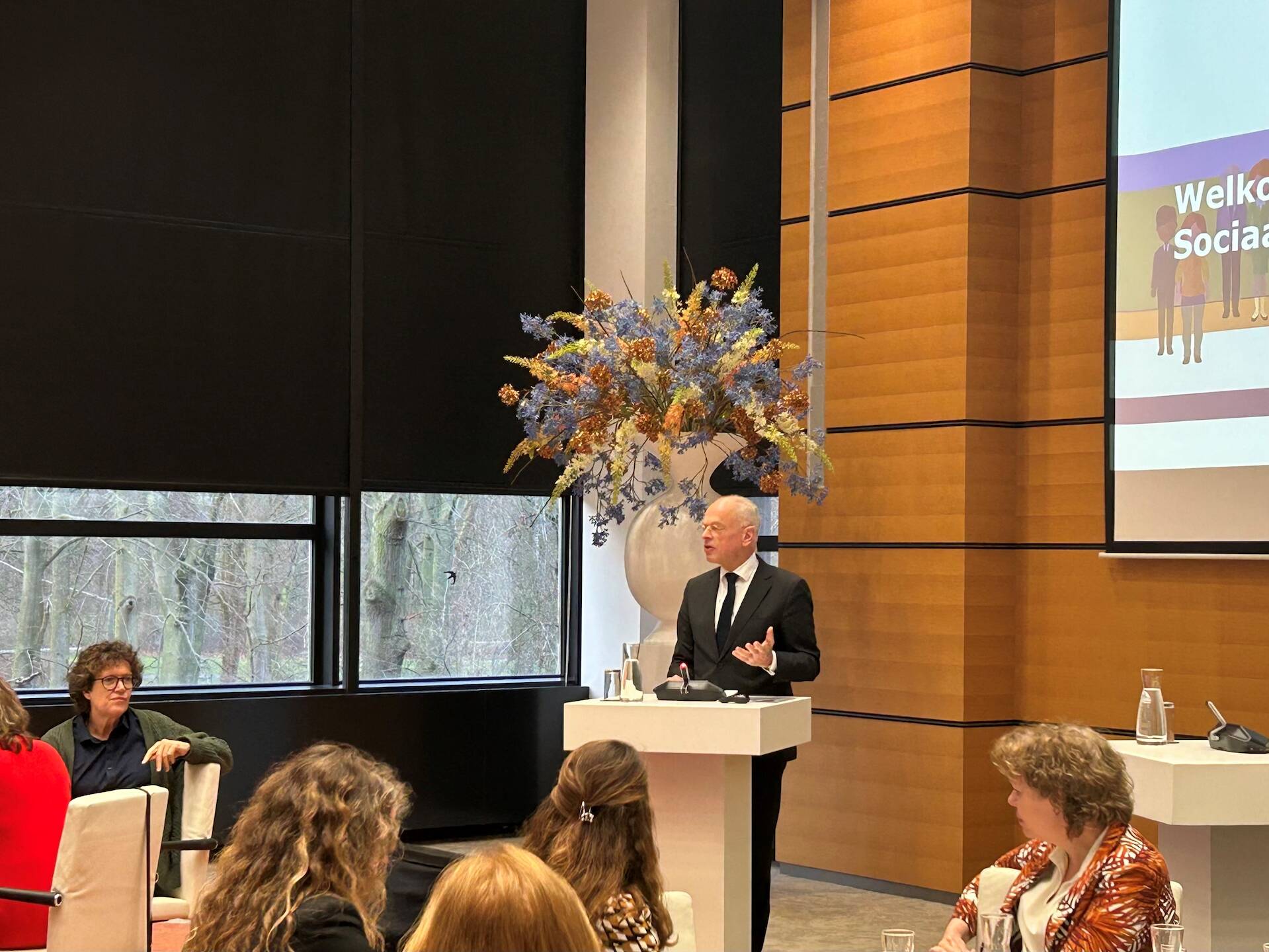 Ingezoomde foto van een zaal met ronde tafels waaraan mensen zitten die luisteren naar een spreker achter het spreekgestoelte. 