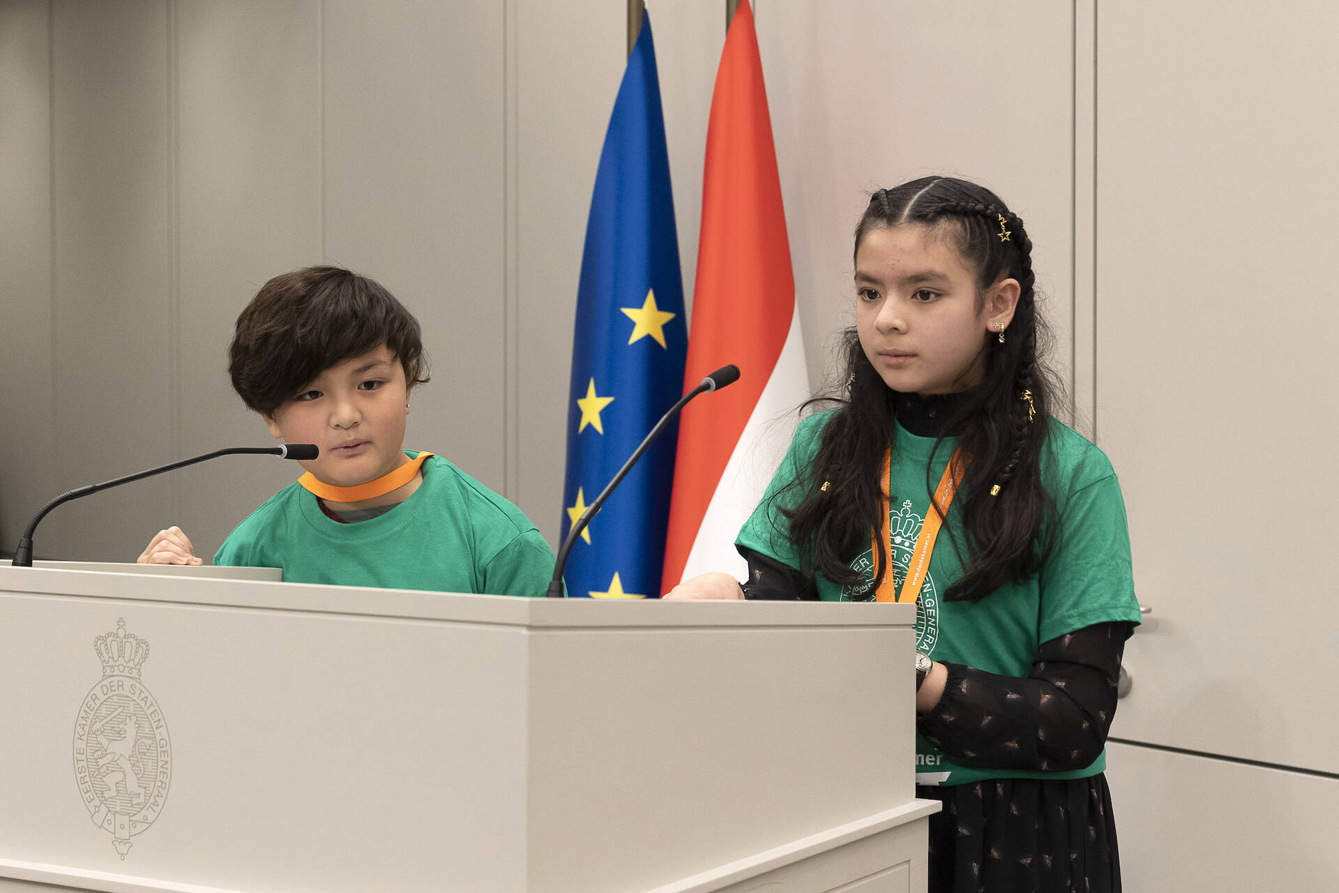 Een jongen en meisje in groene shirts staan achter het spreekgestoelte met microfoons.