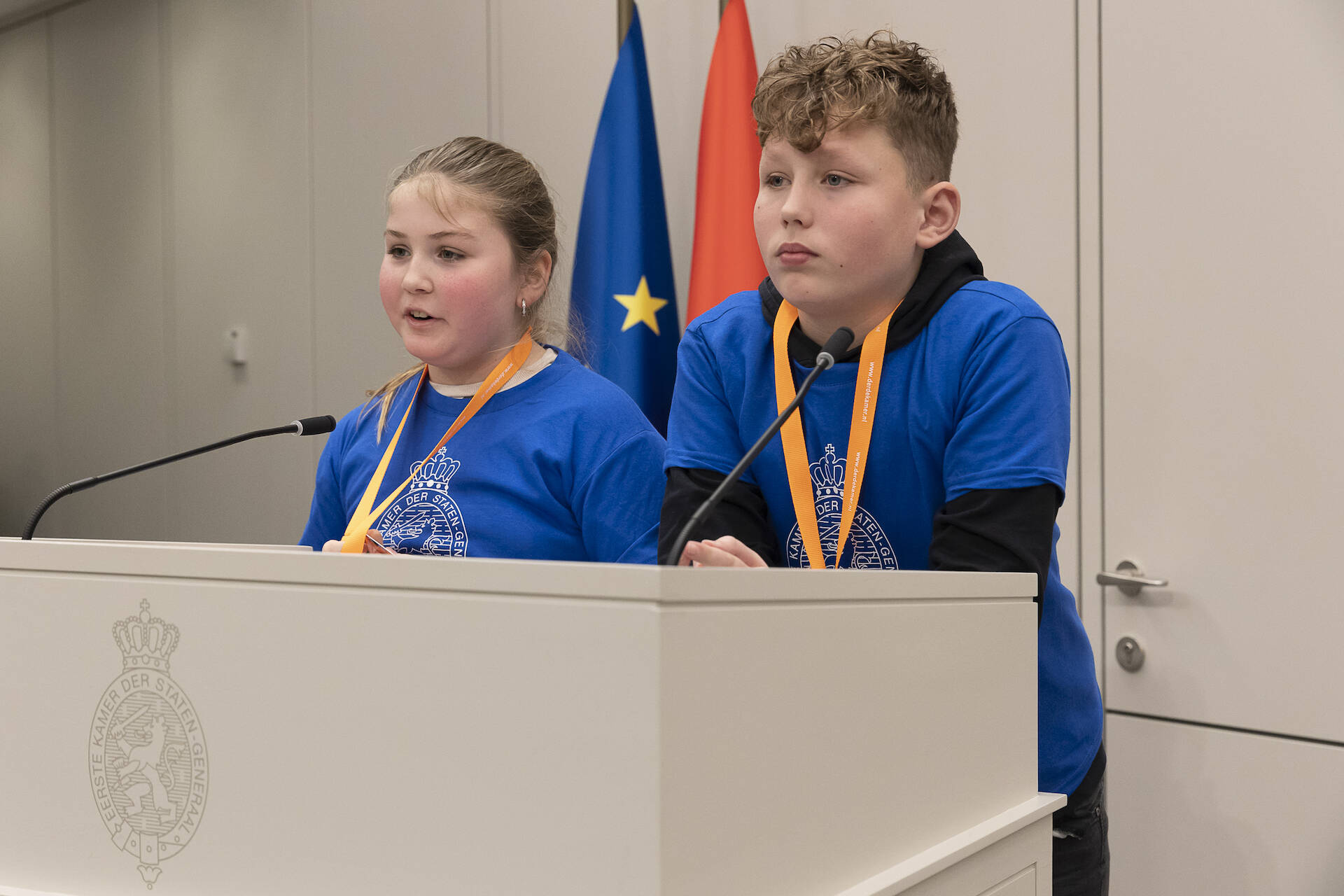 Een jongen en meisje in blauwe shirts staan achter het spreekgestoelte met microfoons