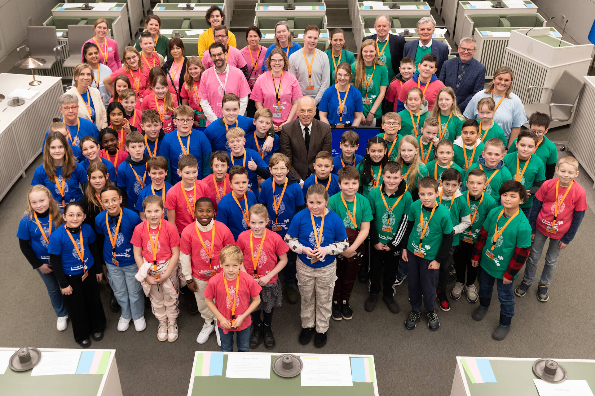 Foto van een grote groep kinderen en enkele volwassenen in de vergaderzaal van de Eerste Kamer. Ze dragen t-shirts in de kleuren roze, groen en blauw.