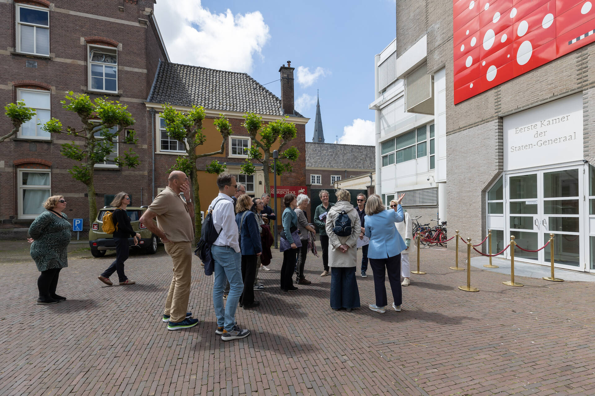 Foto van de gevel van de hoofdingang van de Eerste Kamer met daarvoor een groep mensen die uitleg krijgt over het kunstwerk aan de gevel