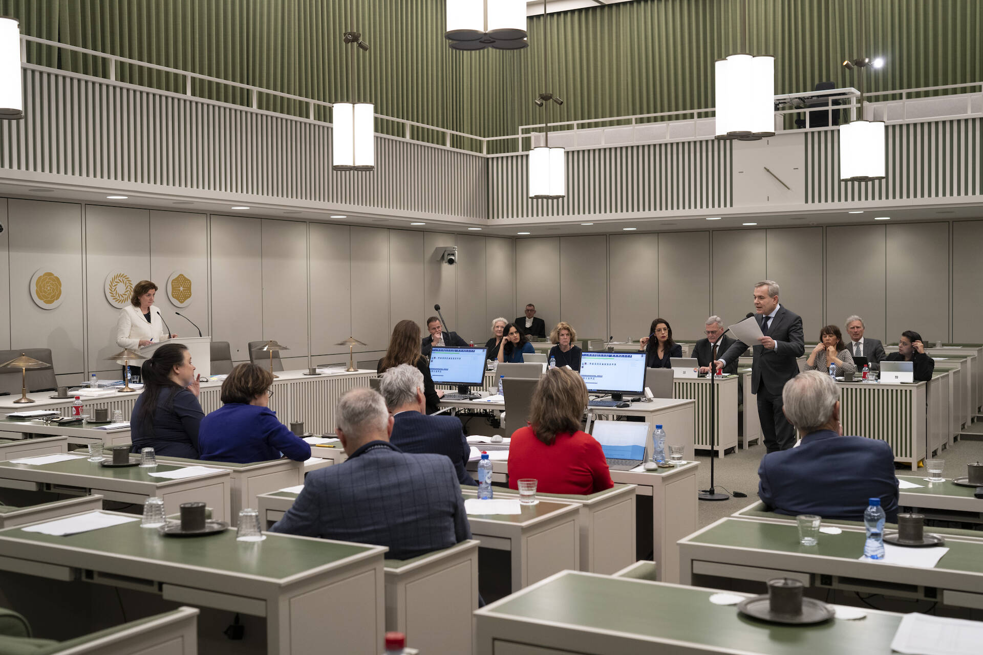 Foto van de plenaire vergaderzaal van de Eerste Kamer: mensen zitten in bankjes, een persoon staat bij de interruptiemicrofoon en een persoon staat achter het spreekgestoelte op de regeringstafel